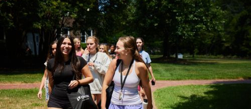 Students interested in medicine are walking and chatting in a park-like setting, surrounded by trees and grass under bright sunlight, creating a cheerful and relaxed atmosphere.