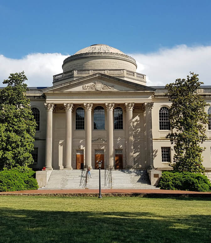 A neoclassical building features a domed roof, tall columns, and three entrance doors. A person walks down the steps amidst surrounding greenery, with a backdrop of a clear blue sky.