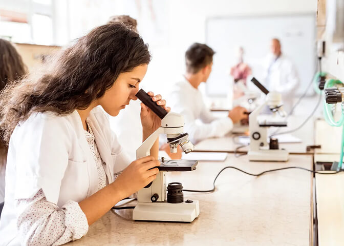 A student looks through a microscope, surrounded by others using microscopes, in a laboratory setting. Whiteboards and lab coats in the background suggest a scientific or educational environment.