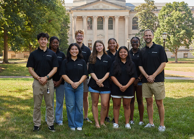 Nine Outset staff members in matching black shirts stand together smiling, outdoors on grass. Behind them is a large, neoclassical building with columns, surrounded by trees.