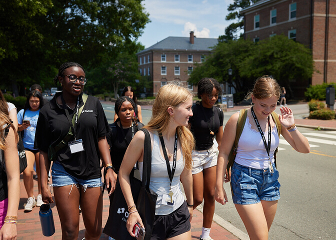 A group of students interested in medicine walk together on a sunny day, wearing casual summer clothing with lanyards, near a brick building and trees, suggesting a campus environment.