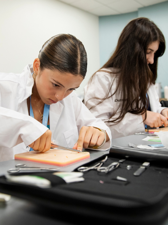 Two students in lab coats practice surgical techniques on simulators, using instruments on a table, within a classroom setting.