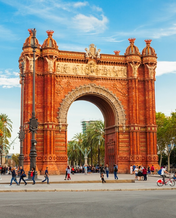 An ornate, red-brick arch stands prominently; people stroll around and gather underneath; palm trees and bright sky create a lively, sunny atmosphere.