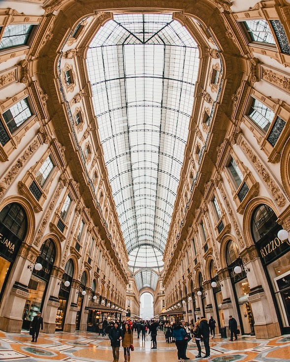 People walk under a grand, glass-vaulted ceiling in an ornate shopping arcade. The scene is flanked by elegant storefronts, including Prada and Rizzoli, with intricate architectural details.