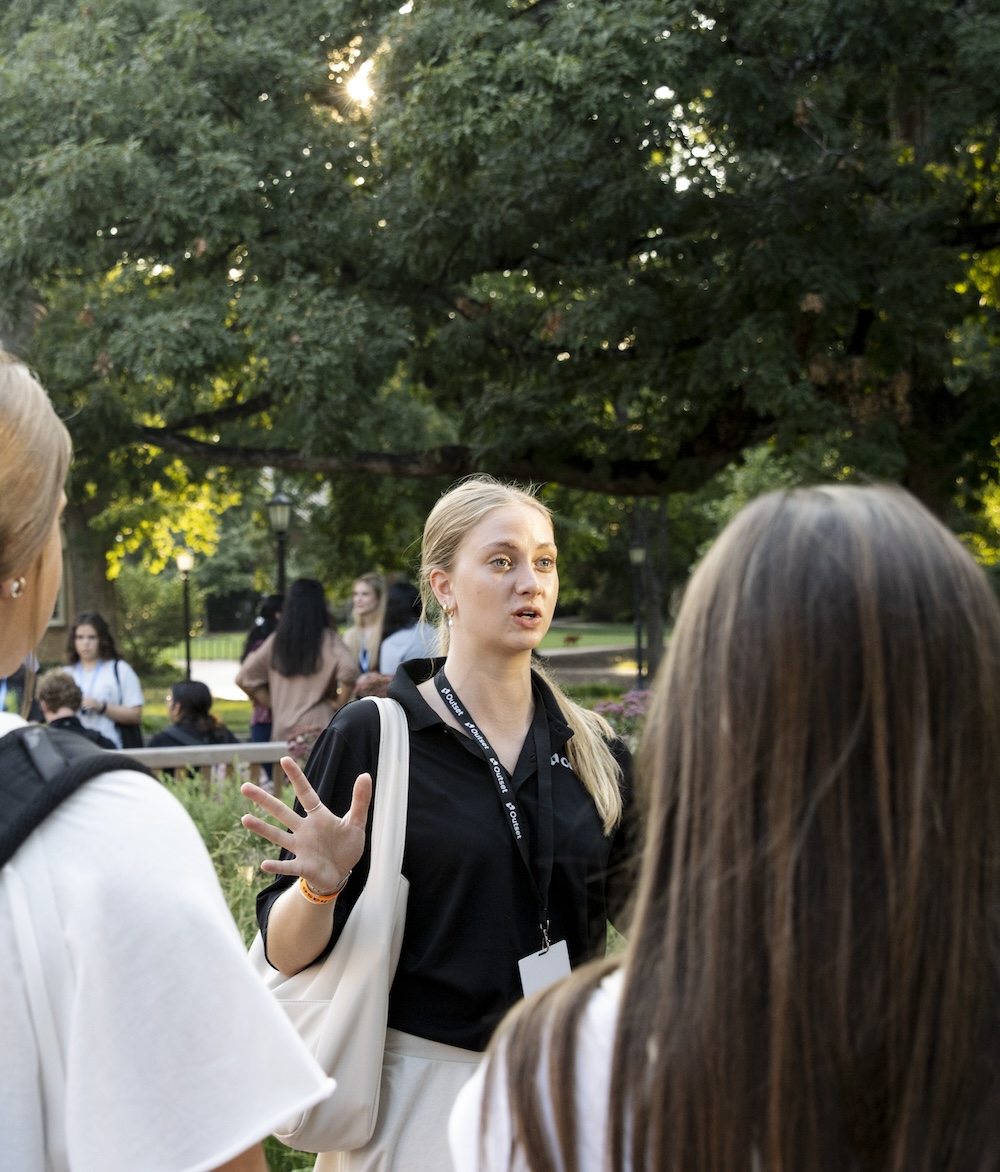 An Outset staff member gestures emphatically while talking to a group, in a sunlit park setting. The atmosphere appears lively with several people and trees in the background.