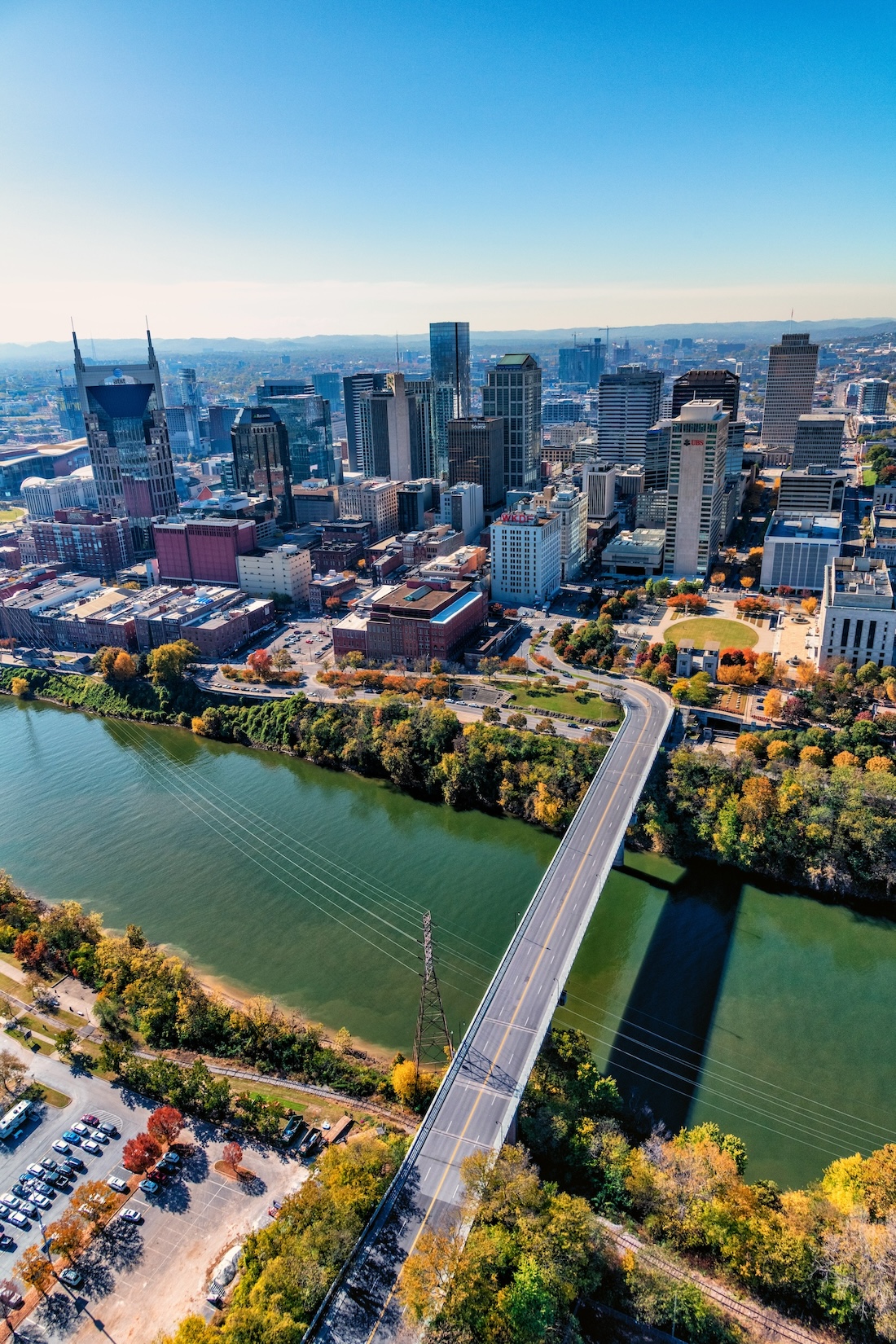 Skyscrapers form a city skyline under a clear sky, with a bridge crossing a river in the foreground. Autumn trees surround the water, creating a colorful contrast between urban and nature.
