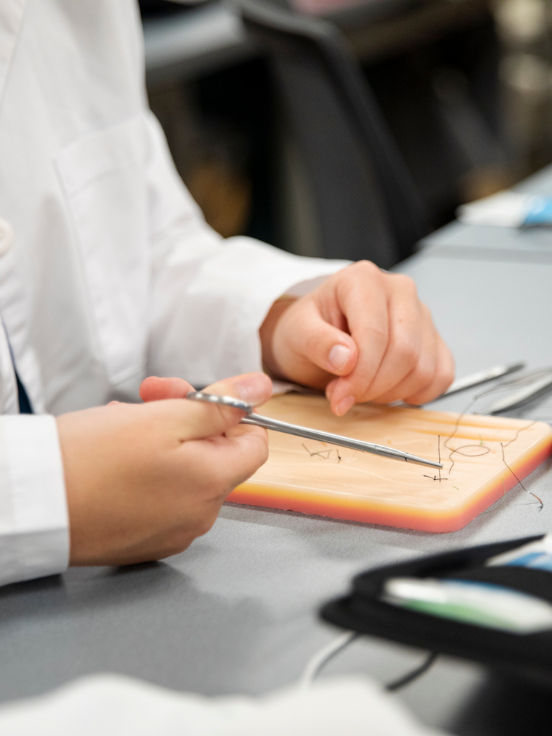 Hands hold surgical tools practicing sutures on a simulation pad. The student wears a white coat, working at a table in a classroom or training environment.